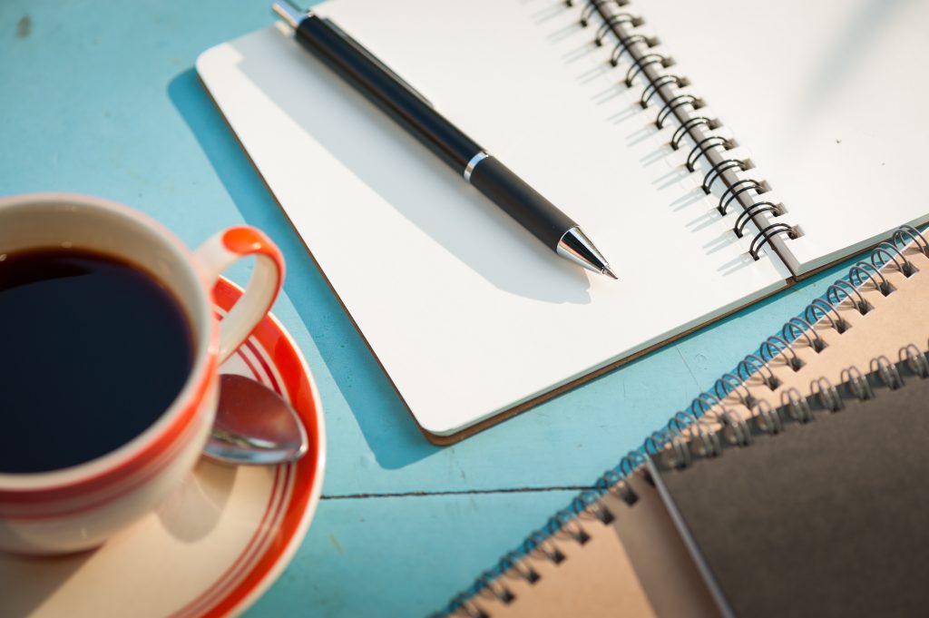 An open notebook, pen, and cup of coffee on a table, set out for reflective practice